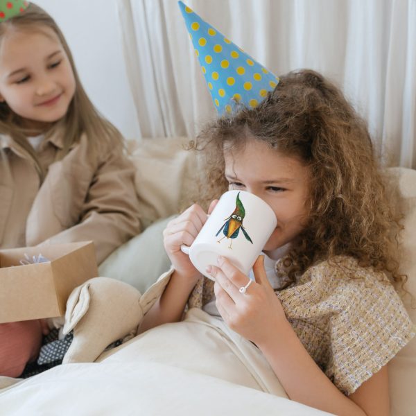 Two little girls with party hats cozy setting one girl drinking from a white ceramic coffee mug featuring a watercolor painting of a whimsical crow called Bubba