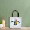 White polyester tote bag featuring a whimsical watercolor painting of a crow the bag is leaning on a green wall