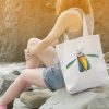 woman on a rocky beach with a a white canvas tote bag featuring a watercolor painting of a whimsical crow