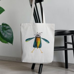 a white canvas tote bag featuring a watercolor painting of a whimsical crow bag is on a chair