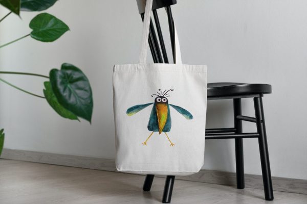 a white canvas tote bag featuring a watercolor painting of a whimsical crow bag is on a chair