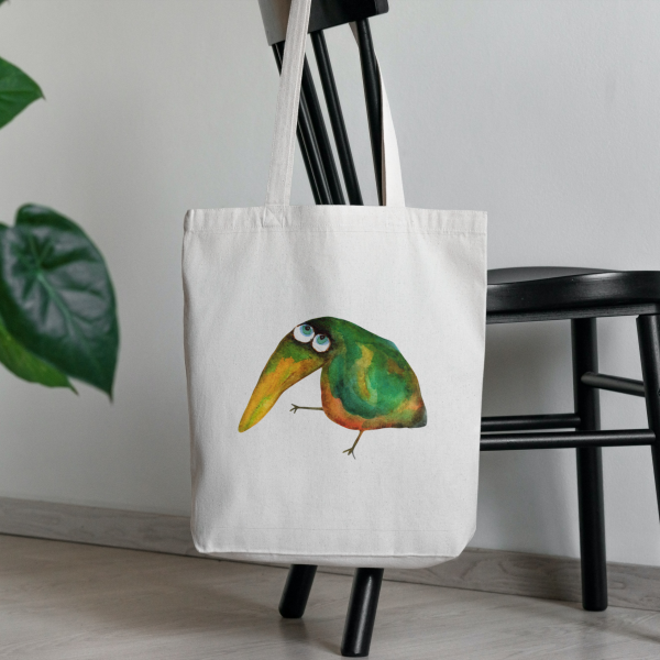 a white canvas tote bag featuring a watercolor painting of a whimsical crow bag is hanging on a chair