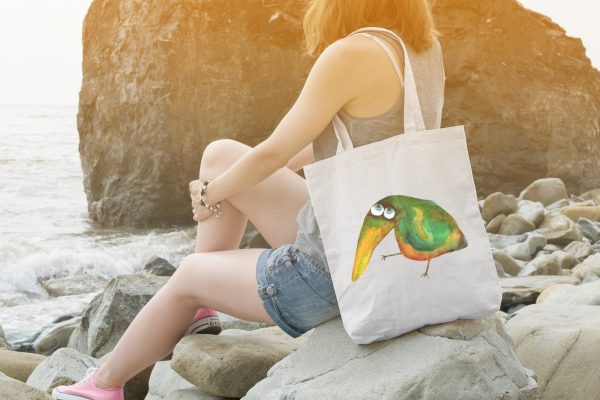girl on a rocky beach with a white canvas tote bag featuring a watercolor painting of a whimsical crow