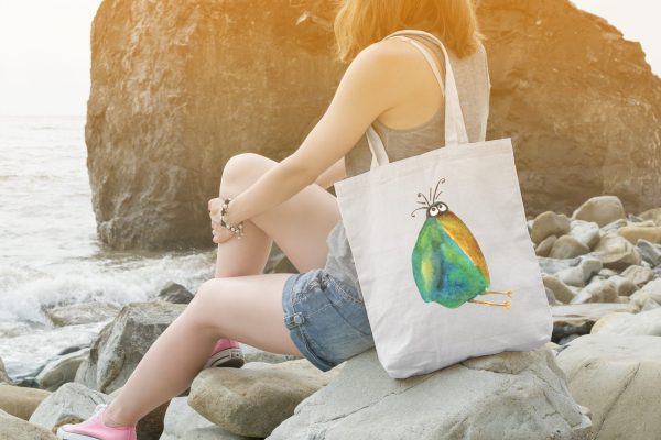 girl on a rocky beach with a white canvas tote bag featuring a watercolor painting of a whimsical crow