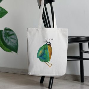 a white canvas tote bag featuring a watercolor painting of a whimsical crow bag is on a chair