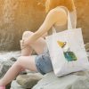 girl on a rocky beach with a white canvas tote bag featuring a watercolor painting of a whimsical crow