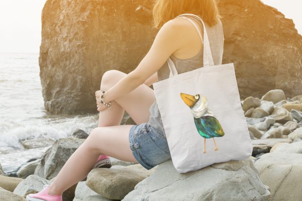 girl on a rocky beach with a white canvas tote bag featuring a watercolor painting of a whimsical crow