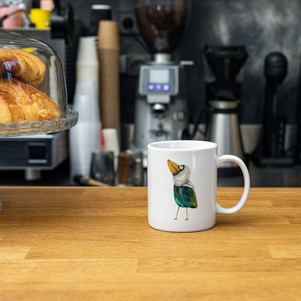 White ceramic coffee mug with a watercolor painting of a whimsical crow called Squawk mug is on a cafe table