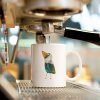 White ceramic coffee mug with a watercolor painting of a whimsical crow called Squawk mug is standing on an espresso machine