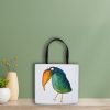 white polyester tote bag leaning against a wall. The tote bag features a watercolor illustration of a whimsical crow