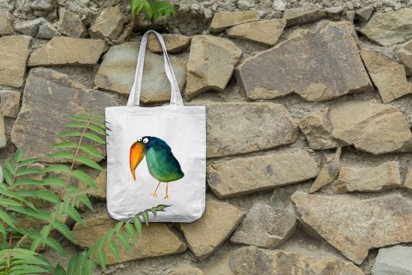 a white canvas tote bag featuring a watercolor painting of a whimsical crow bag is hanging on a wall