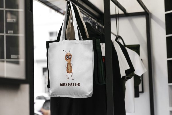 white canvas tote bag with a watercolor painting of a whimsical ant bag is hanging on a rack