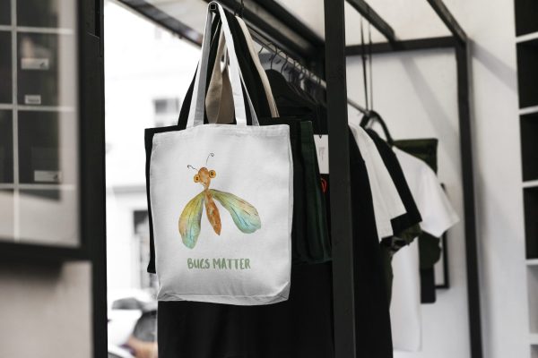 white canvas tote bag with a watercolor painting of a whimsical green lacewing bag is hanging on a rack