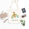 white canvas tote bag with a watercolor painting of a whimsical green lacewing bag is on the floor
